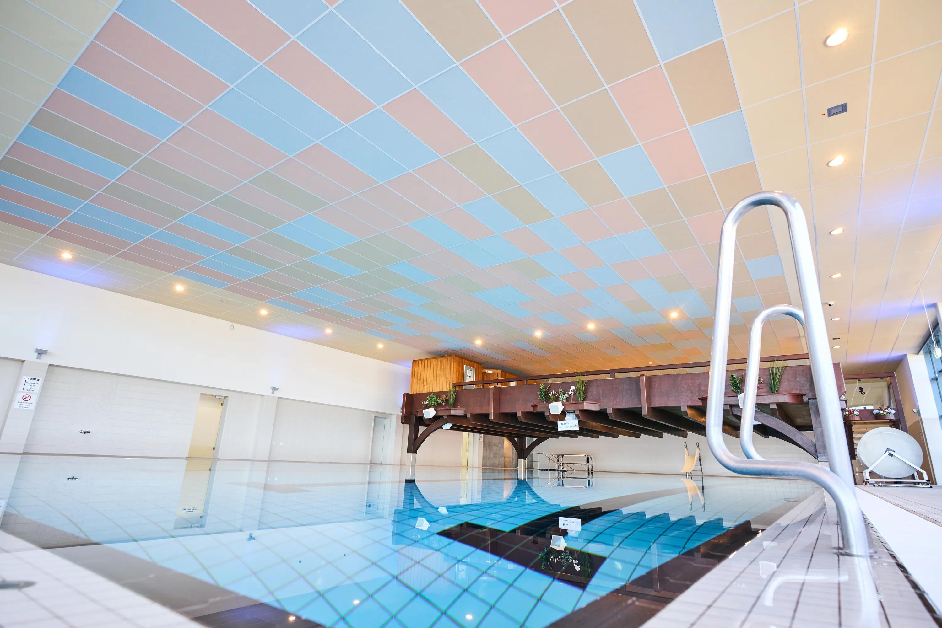 Indoor swimming pool with a pastel-coloured acoustic tile ceiling, calm water surface, and a wooden poolside deck overlooking the pool area.