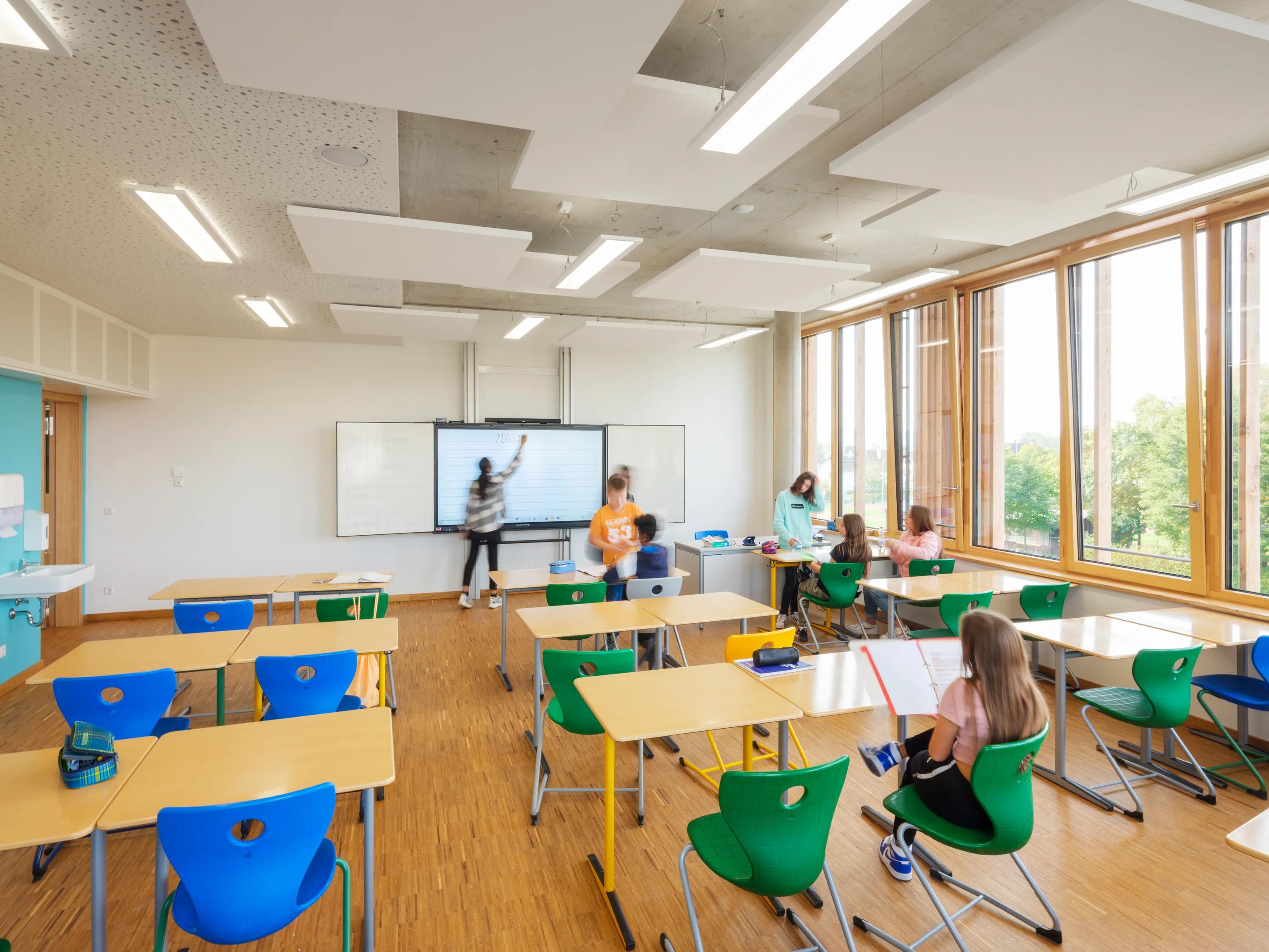 A contemporary classroom featuring white rectangular acoustic ceiling rafts hung beneath an exposed concrete soffit, with students working at desks near tall timber-framed glazing