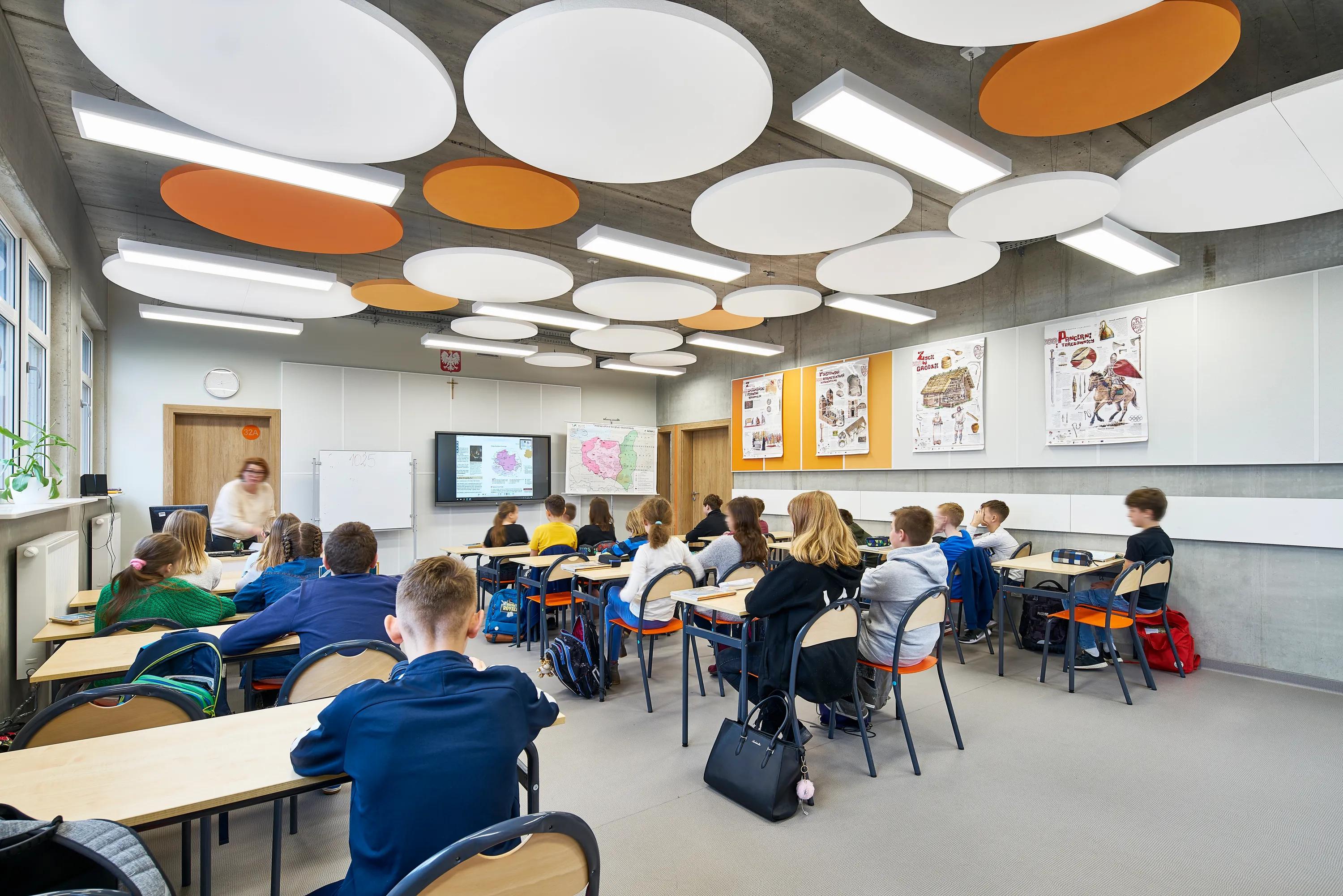 A modern classroom featuring circular acoustic ceiling islands in white and orange, suspended from an exposed soffit, with students seated at desks facing an interactive screen.