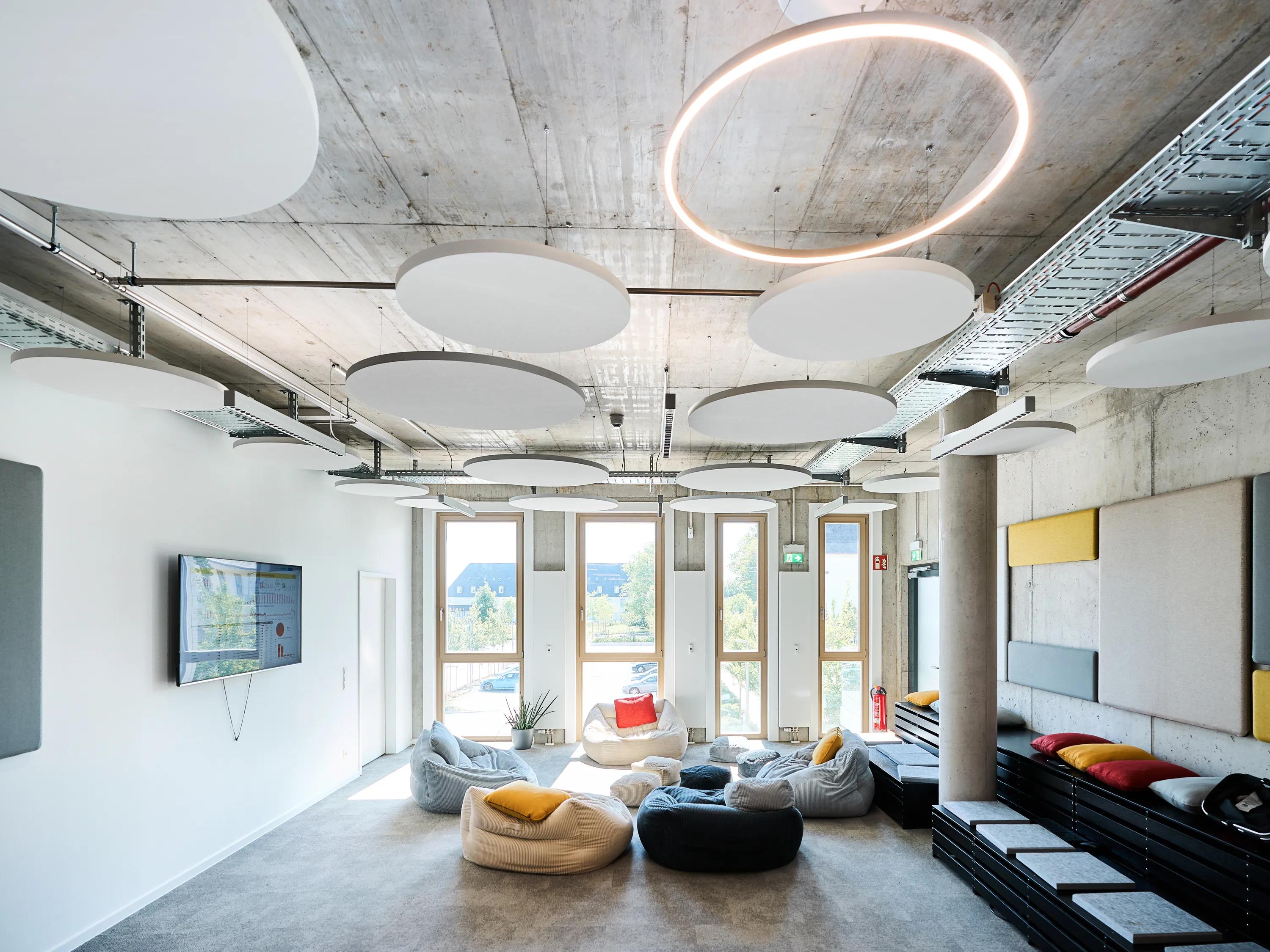 Modern breakout room featuring round acoustic islands on an exposed concrete ceiling, soft seating areas, tiered platforms, and tall windows providing natural lightModern breakout room featuring round acoustic islands on an exposed concrete ceiling, soft seating areas, tiered platforms, and tall windows providing natural light