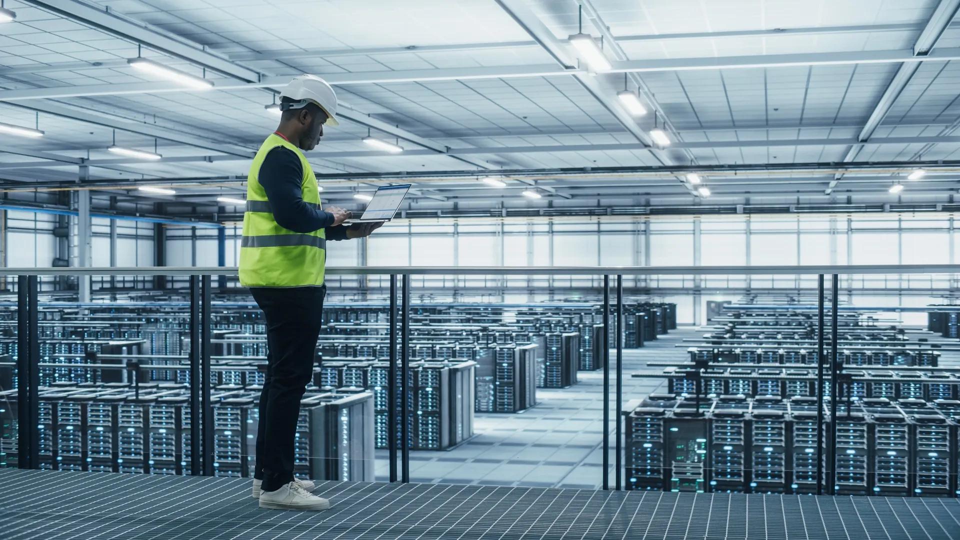 Ein großes Rechenzentrum mit Reihen beleuchteter Server-Racks unter einer abgehängten Akustik-Rasterdecke, die darauf ausgelegt ist, die Geräusche von Kühlsystemen und Geräten zu dämpfen. Ein Techniker in einer Warnweste und mit Schutzhelm steht auf einem erhöhten Übergang und überwacht die Systeme auf einem Laptop.