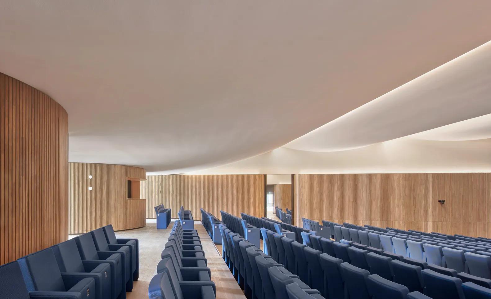 A contemporary auditorium featuring a seamless, wave-like acoustic ceiling above timber wall panels and rows of blue upholstered seats