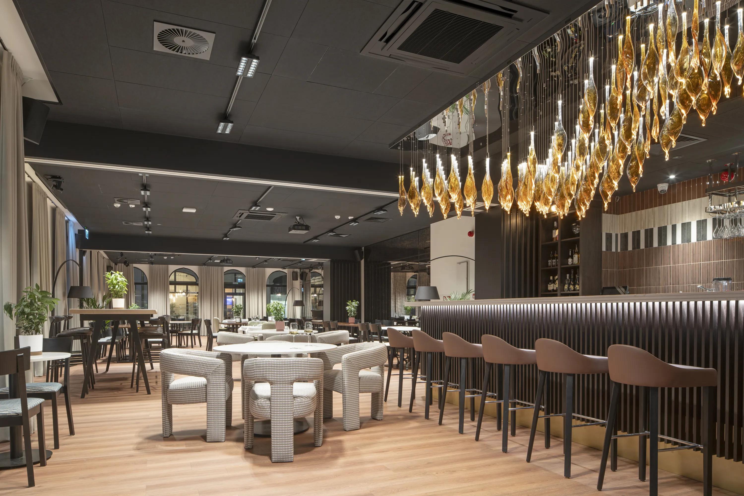 Restaurant interior with dark acoustic ceiling tiles, bar seating, dining tables, and decorative pendant lighting suspended above the bar counter.