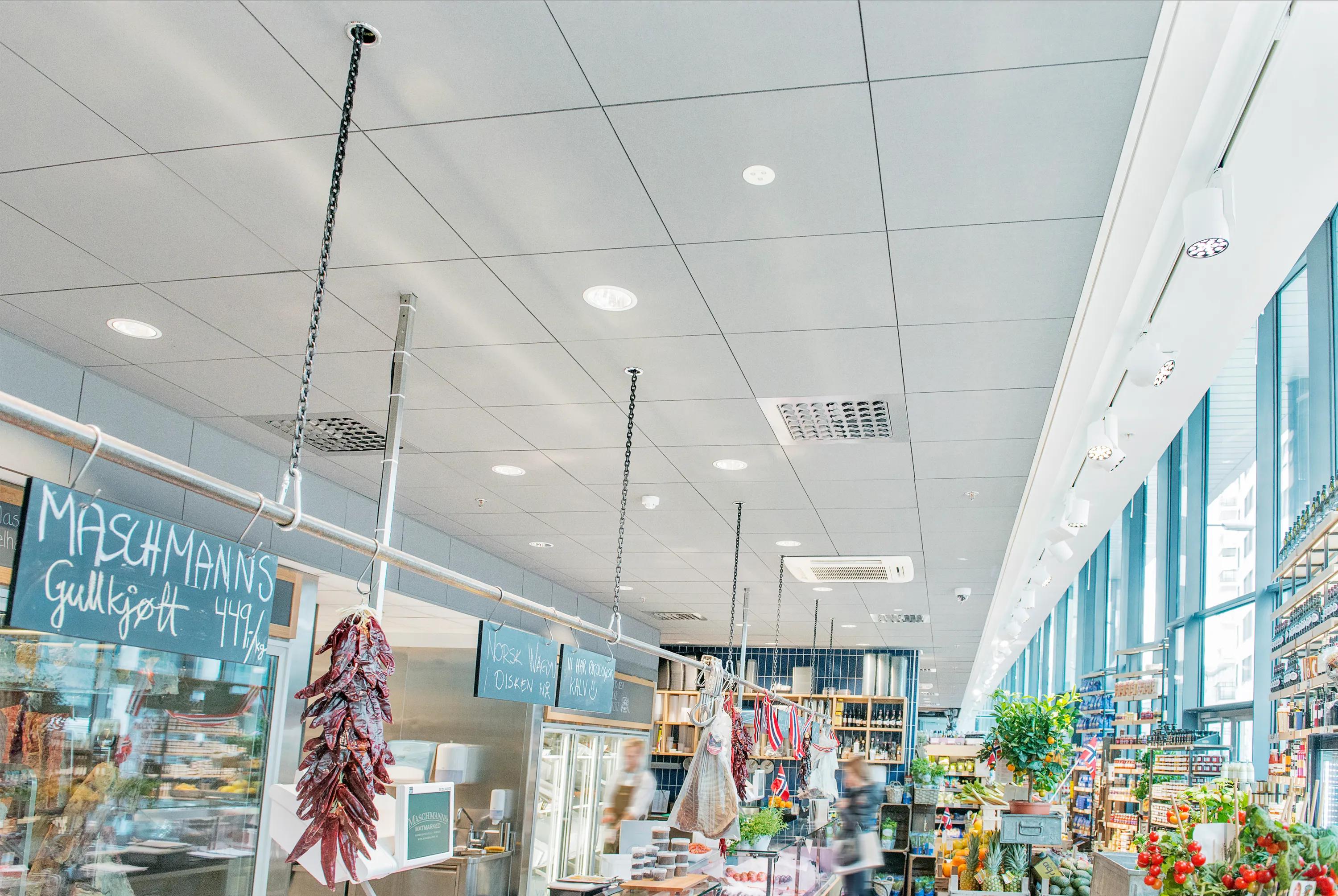 Food court interior with grey acoustic ceiling tiles, pendant lighting, open food counter, and dining area with tables and seated guests in a bright, busy setting.