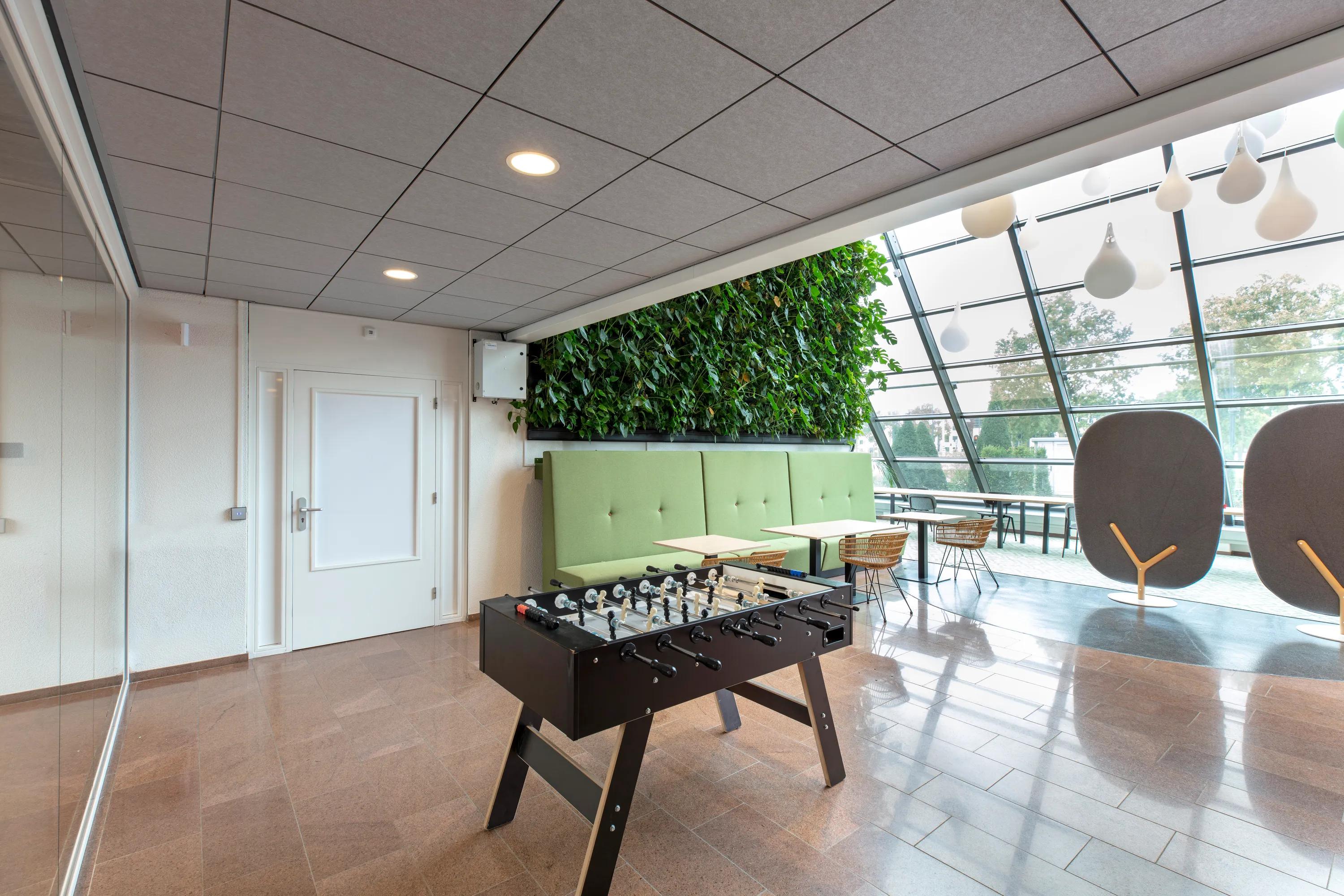 Office breakout area with grey acoustic ceiling tiles, foosball table, green seating, a living plant wall, and large windows bringing in natural daylight.