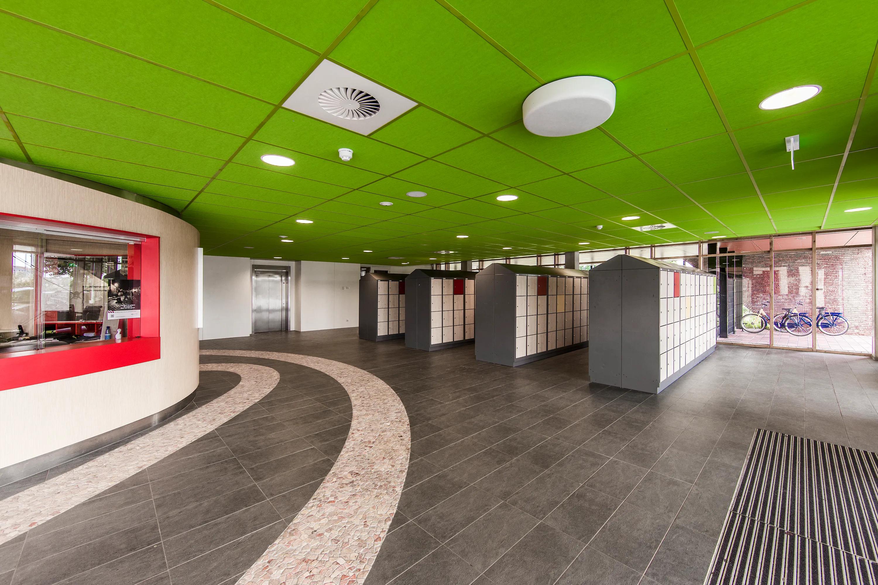 Modern lobby with a green ceiling, red accents, and a curved pathway. Glass doors, elevators, and bike racks are visible in the background.