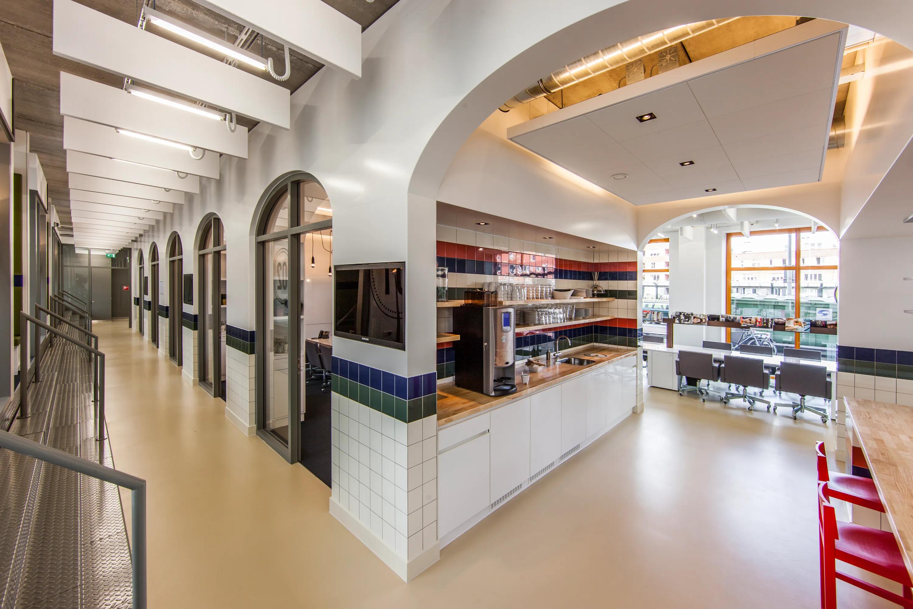 Office corridor and kitchen area with arched openings, white acoustic ceiling baffles along the hallway, and a suspended acoustic ceiling island above the kitchenette.