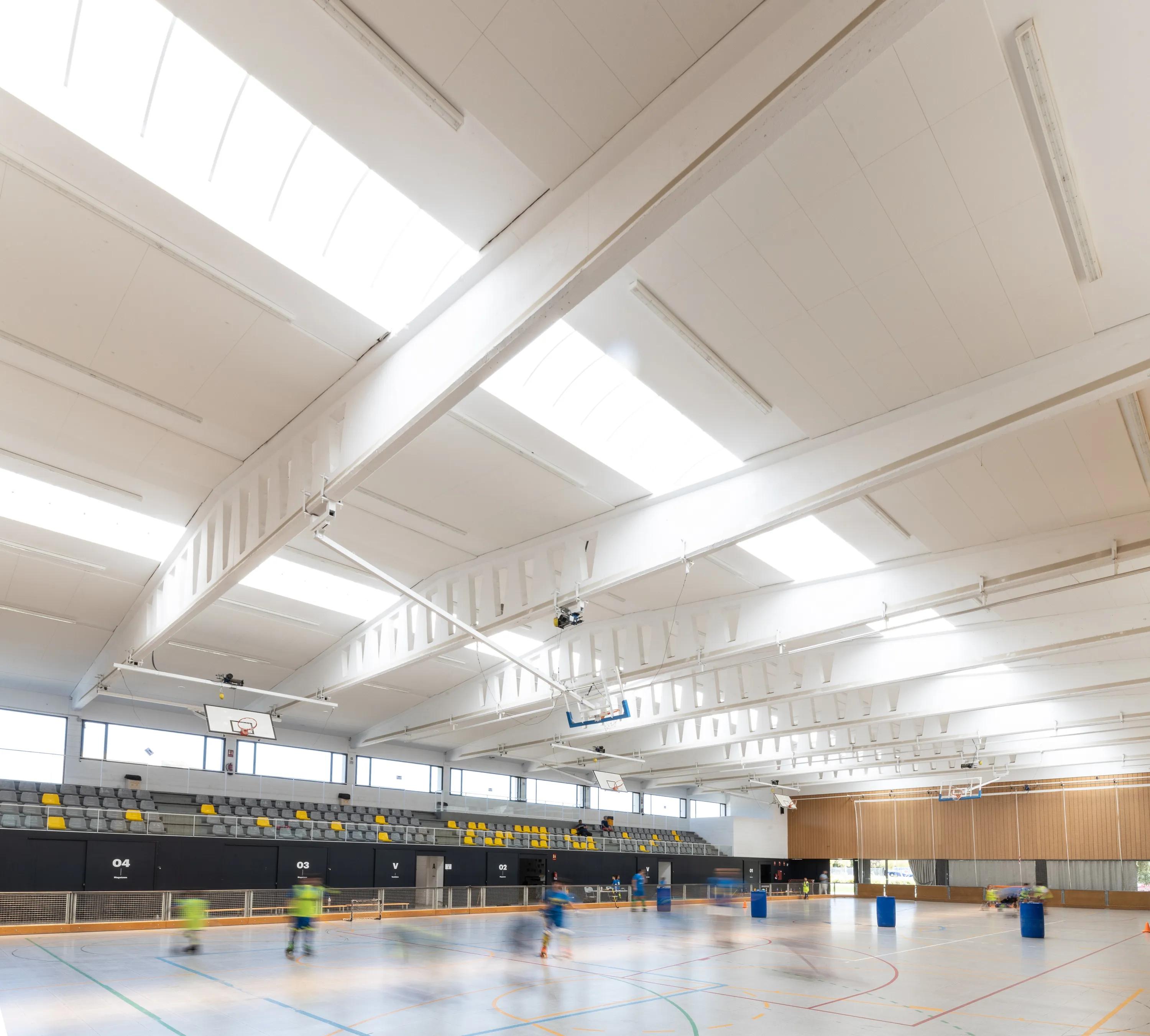 A large sports hall featuring white acoustic ceiling tiles installed between exposed steel beams, with rows of spectator seating and children playing on the multi-purpose court below.