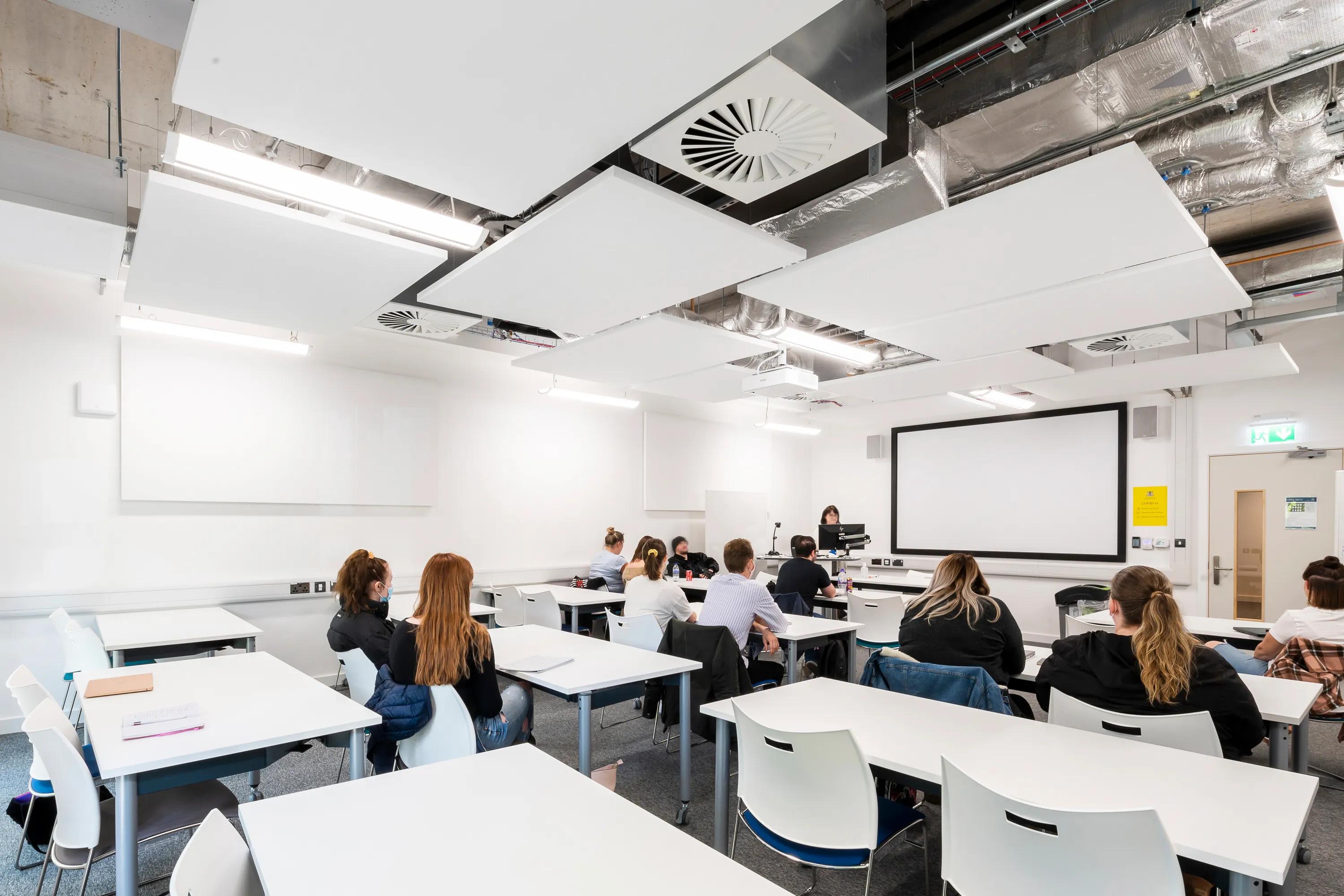 A modern classroom with students seated at white desks beneath suspended white acoustic ceiling rafts and exposed ductwork