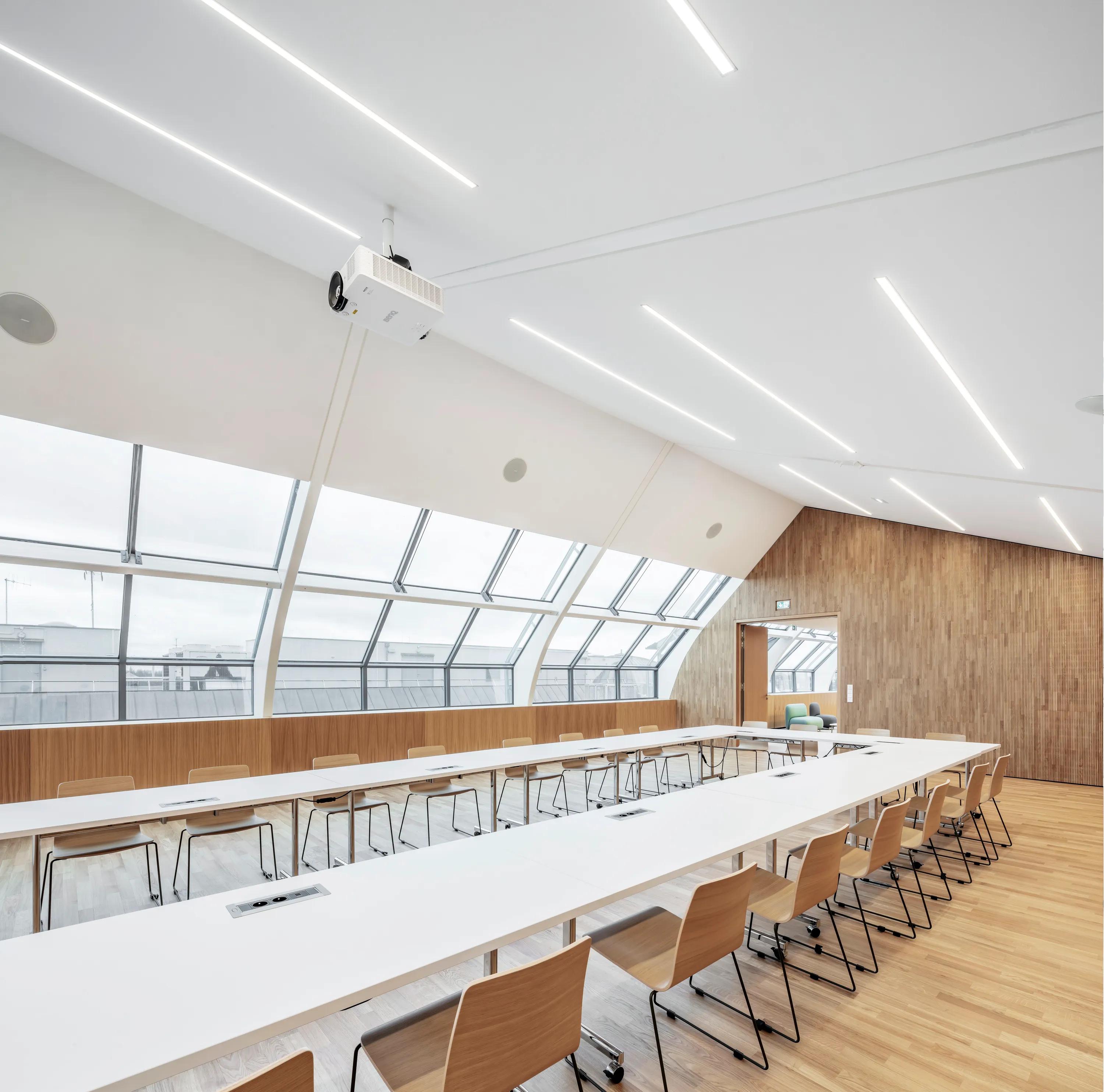 Conference room with a smooth monolithic acoustic ceiling, linear integrated lighting, large skylight windows, and U-shaped tables with wooden chairs.