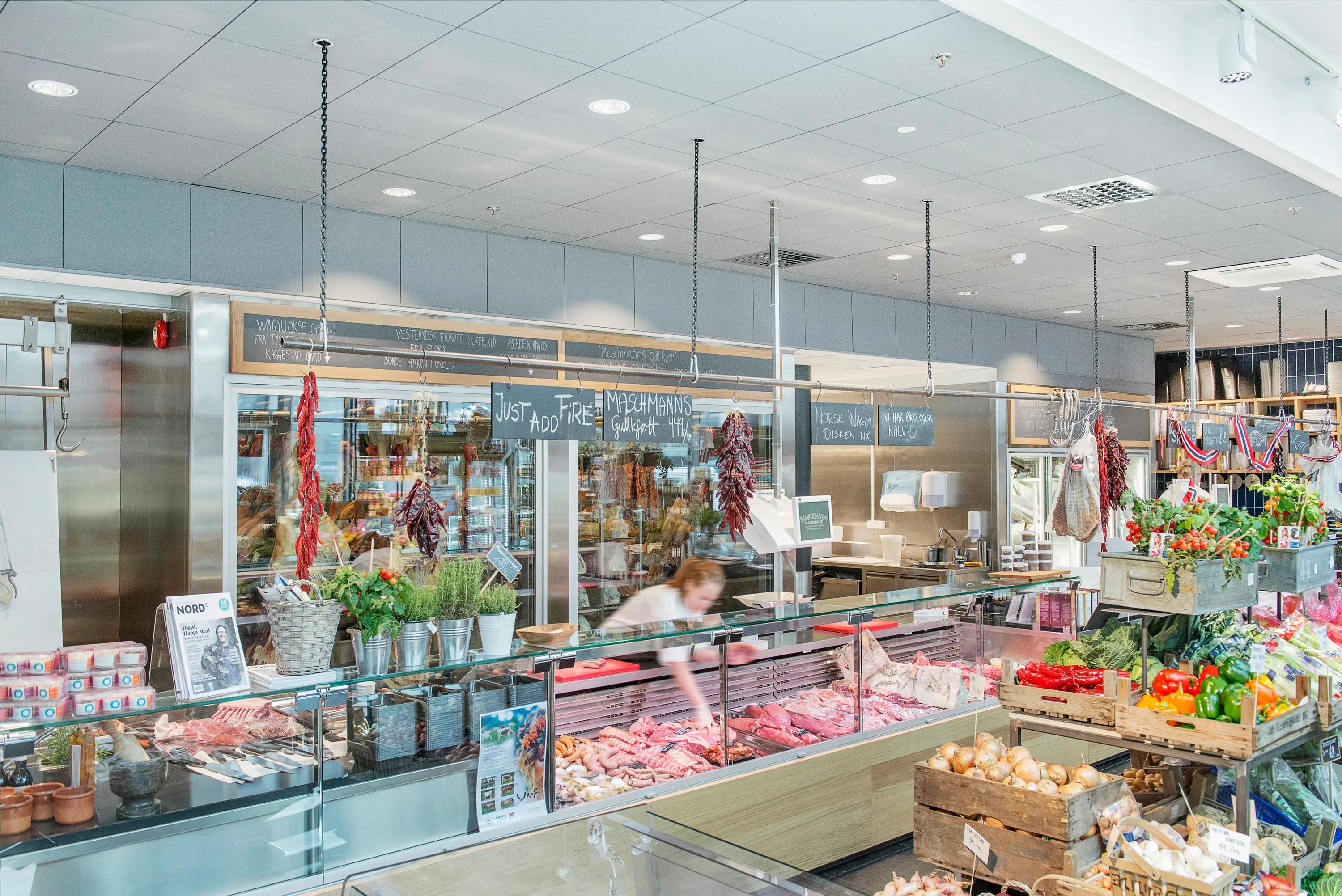 A vibrant indoor food market featuring white acoustic ceiling tiles, recessed lighting, fresh produce stands, and glass-fronted meat counters decorated with herbs and hanging peppers.