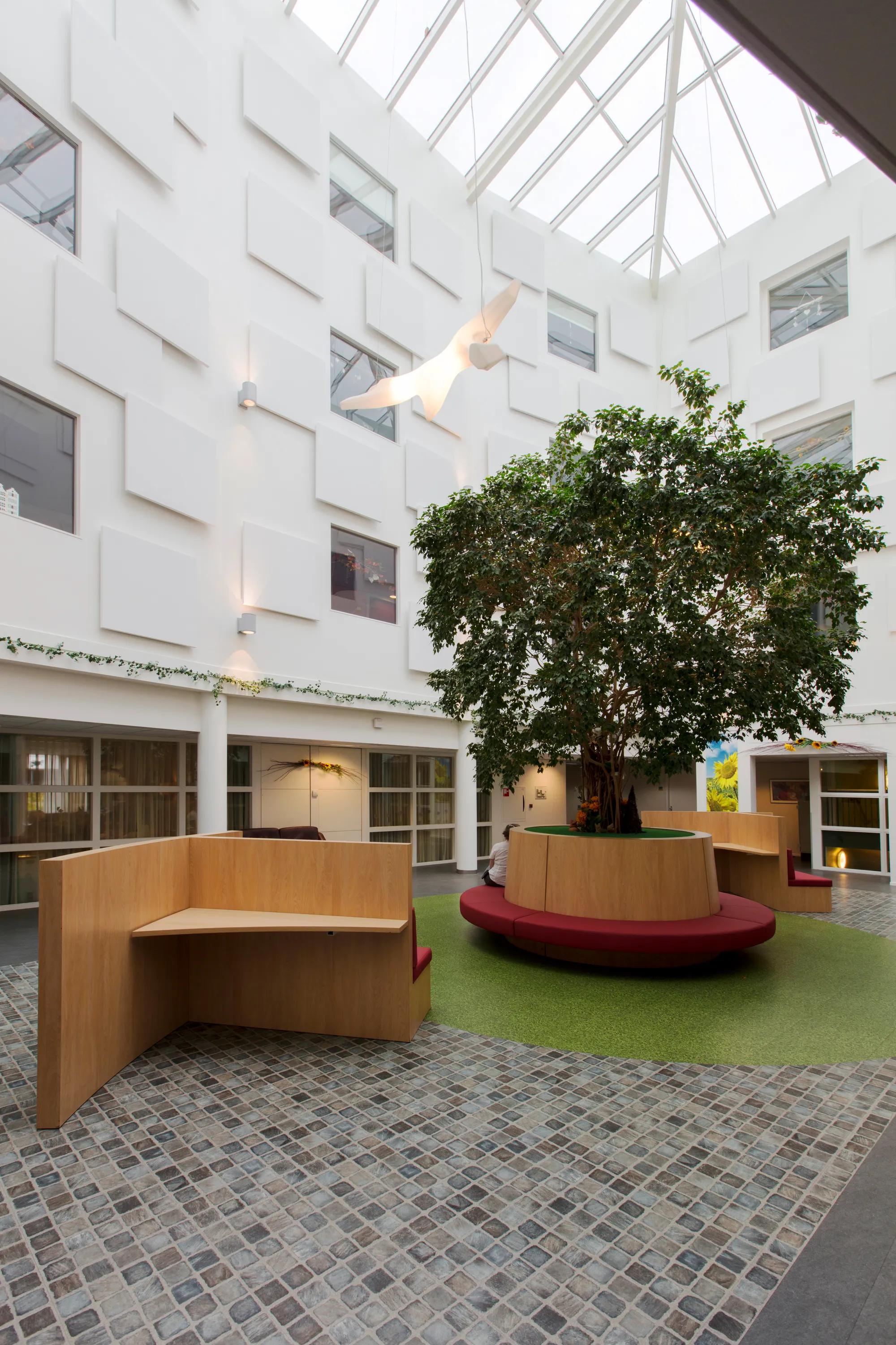 A bright multi-storey atrium featuring staggered white acoustic wall panels and a skylight roof, with seating positioned around a central tree.