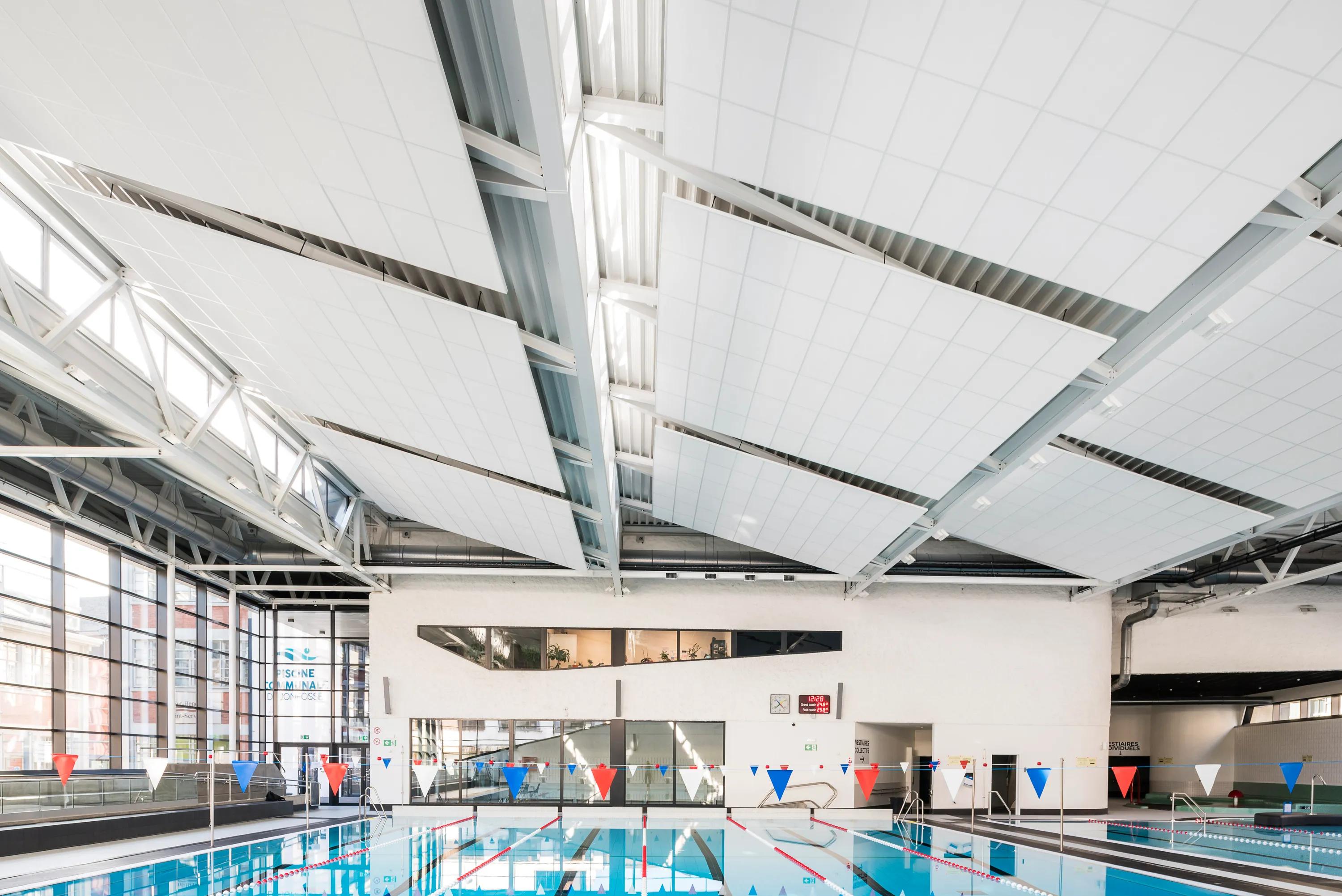 Indoor swimming pool with suspended white acoustic ceiling rafts mounted between exposed structural beams, above a lane pool with flags, large windows, and a bright, open pool hall.