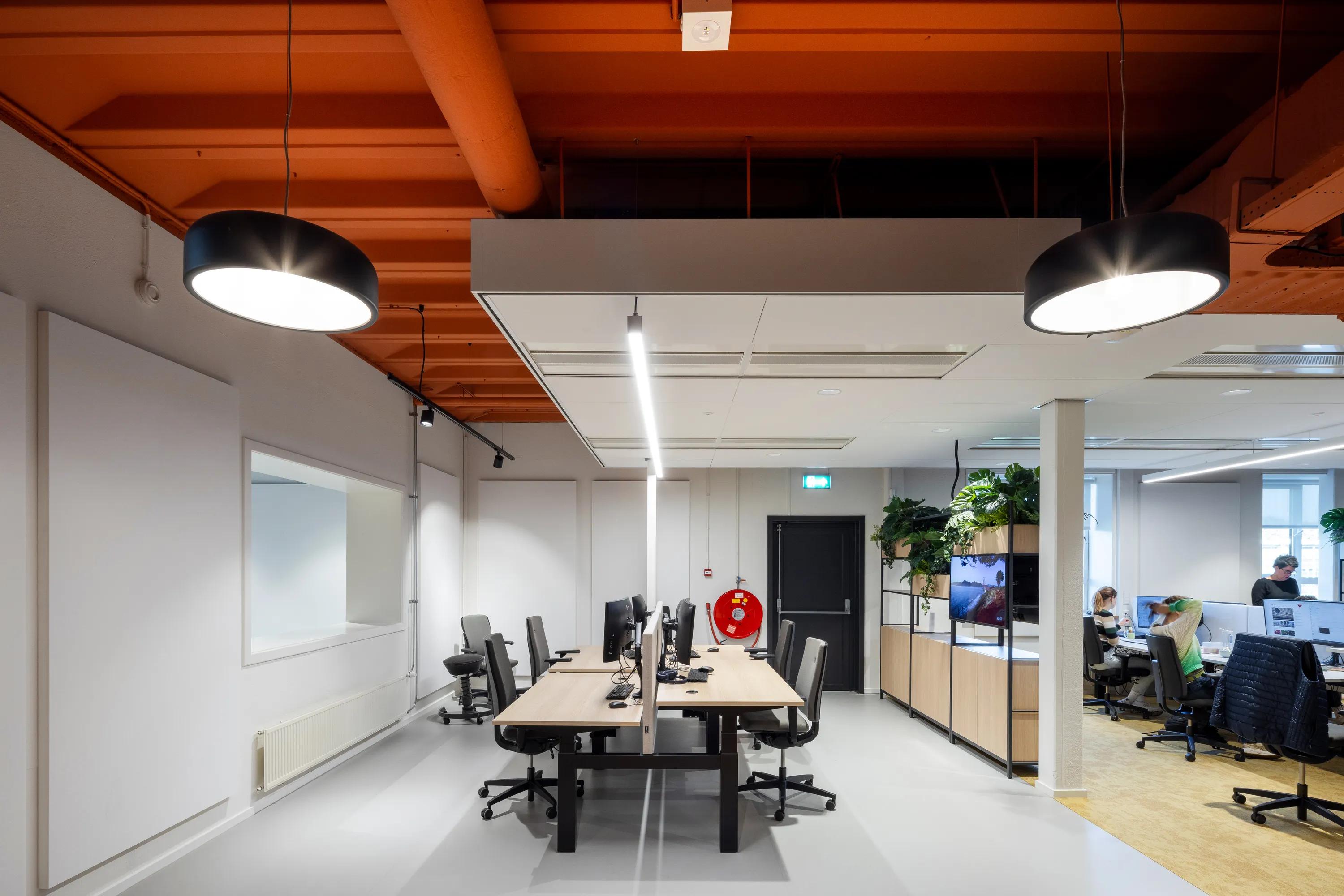 Modern office space with workstations, computers, and plants. Orange ceiling with black pendant lights. People working in the background.