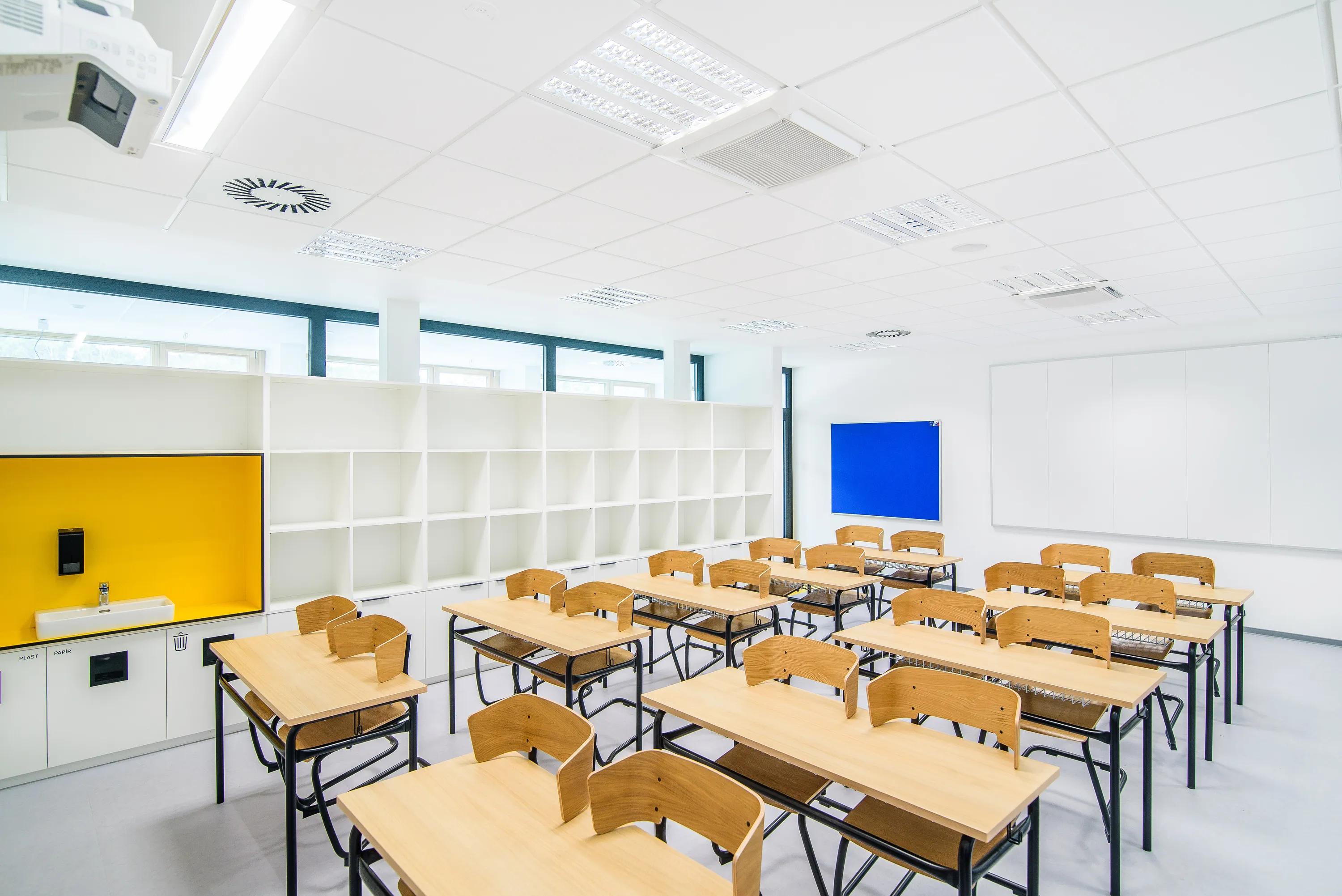 Classroom with white acoustic ceiling tiles, rows of wooden desks and chairs, storage shelving, and a teaching wall with whiteboard and coloured panels.