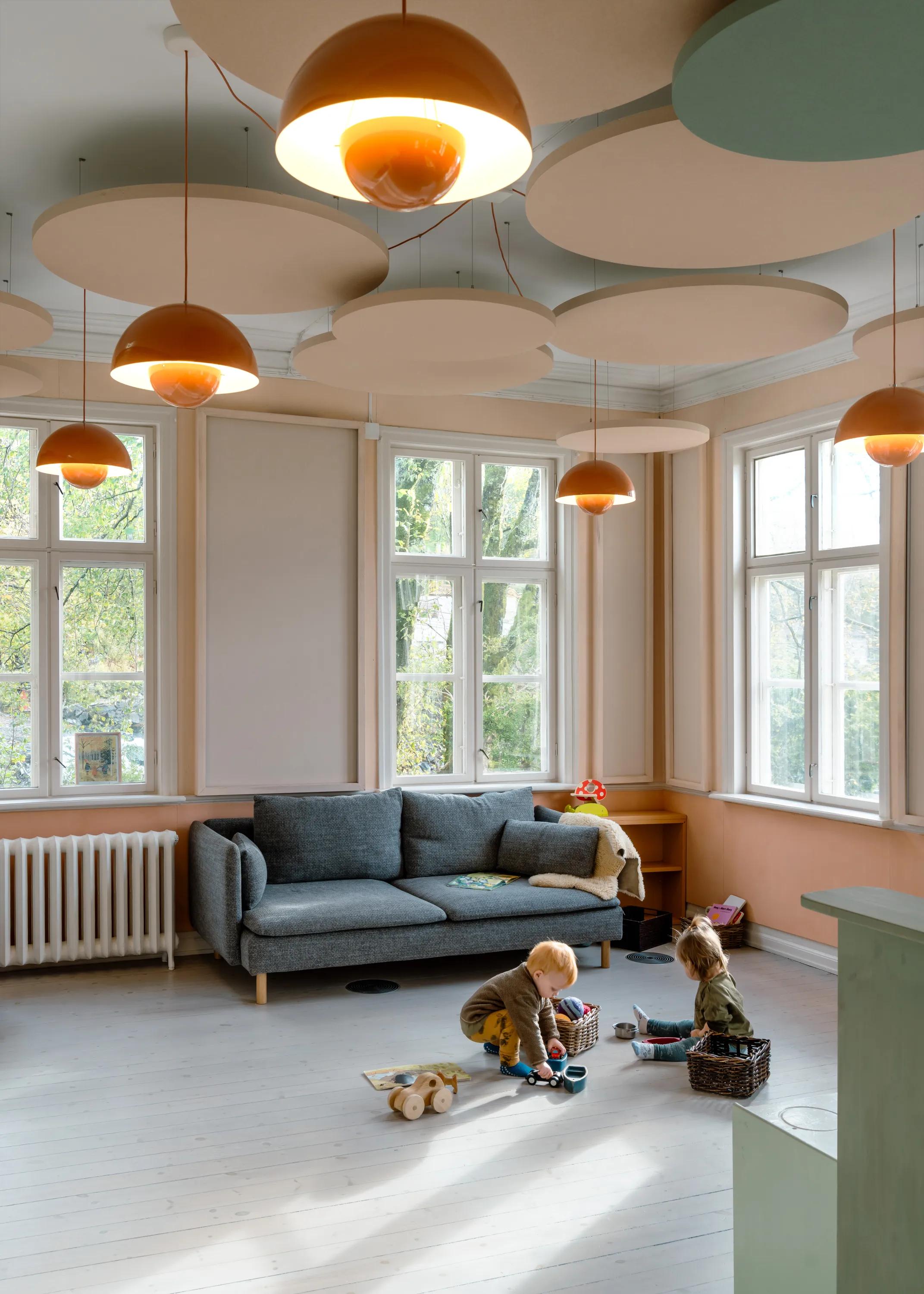 A bright children's playroom featuring suspended circular acoustic ceiling islands and orange pendant lights, with large windows, a grey sofa, and two toddlers playing on the wooden floor.