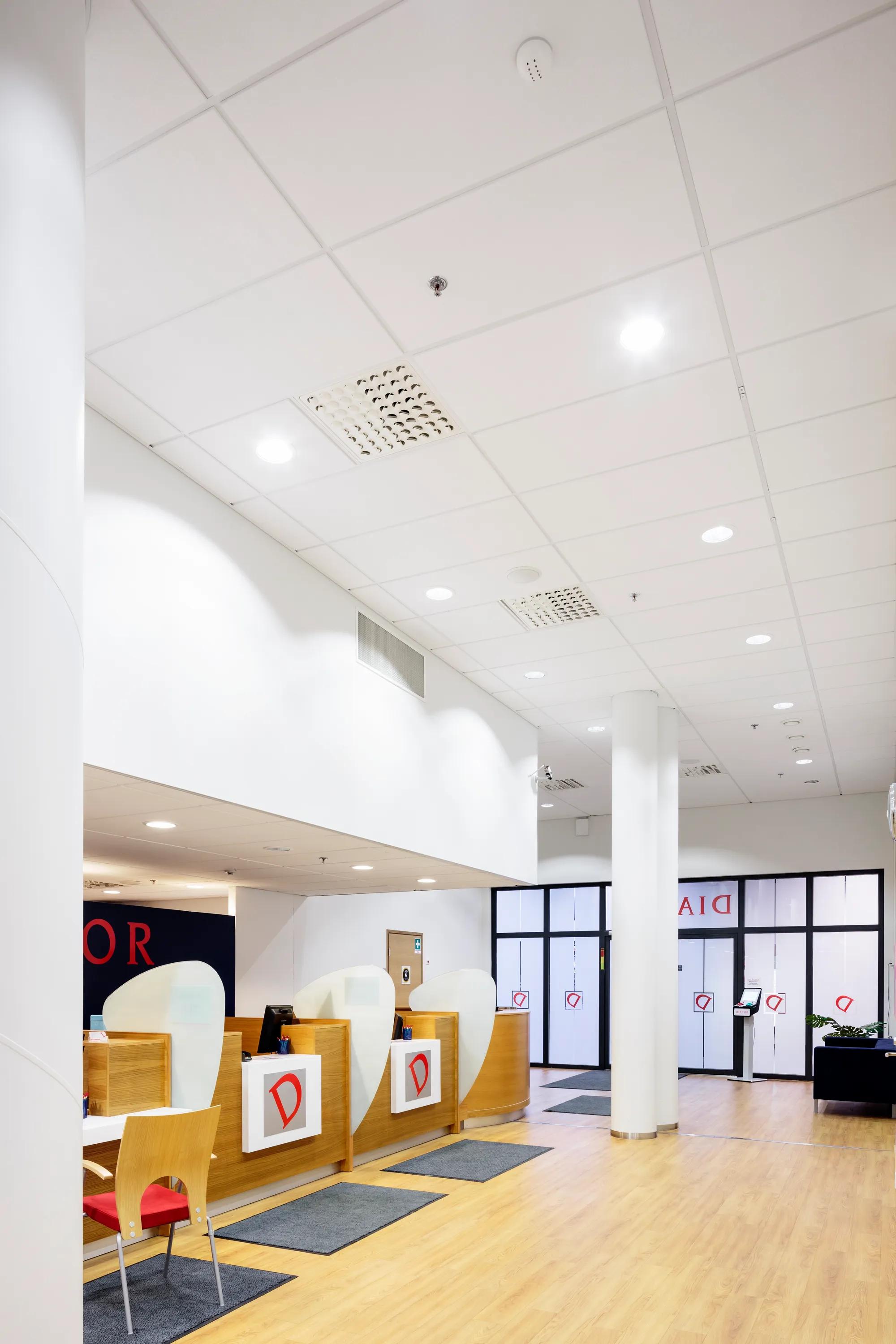 A bright healthcare reception area with wood-panelled service counters, red signage, white acoustic ceiling tiles and recessed downlights.