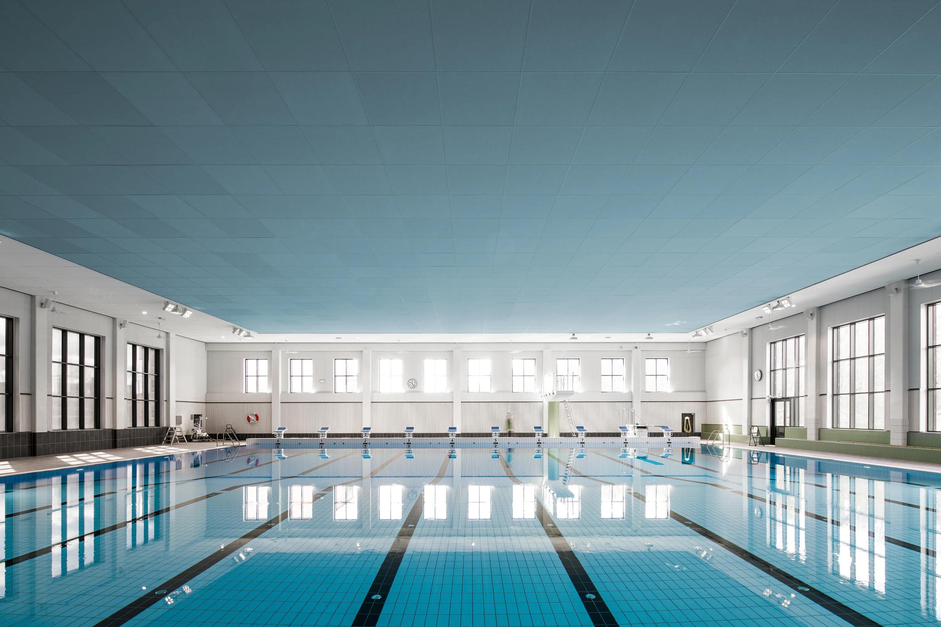 Inomhuspool med en lång bassäng, stora sidofönster, geometriska väggabsorbenter och ett vitt, linjärt akustiskt undertak med integrerad belysning som speglas i vattnet.
