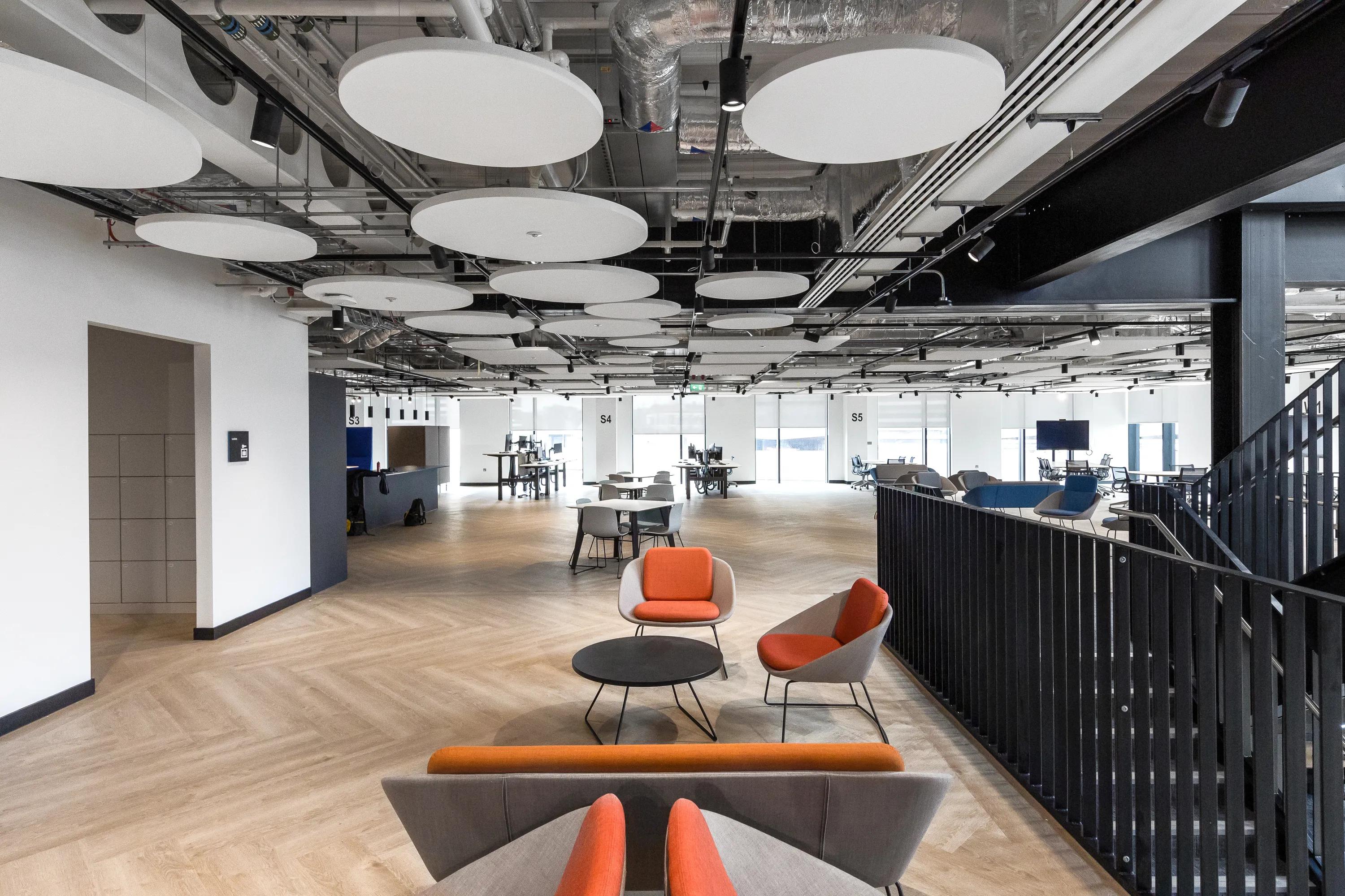 Modern office space with an office ceiling composed of floating circular ceiling panels, open seating area featuring orange and gray chairs, large windows, and wooden flooring.